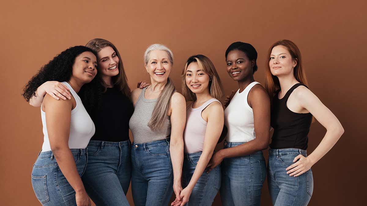 Six women of differing ages and races stand with their arms around each other.