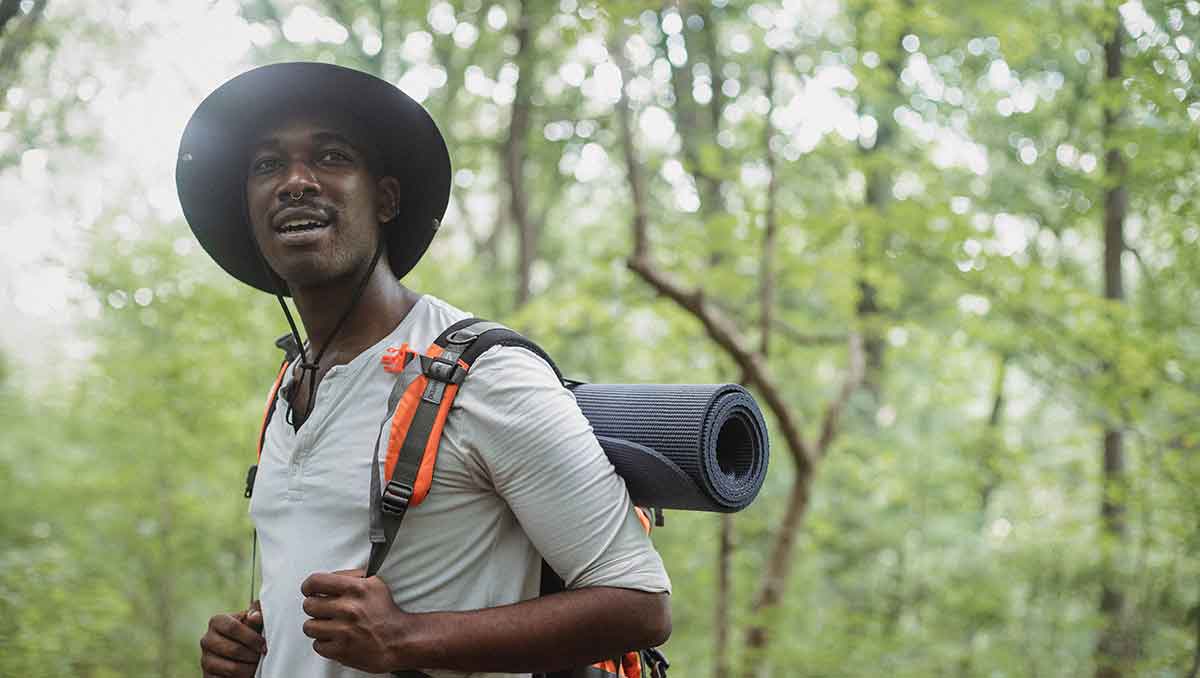 Young Black man wearing a hat and holding the straps on his backpack. He's on an overnight hiking trip in the woods.