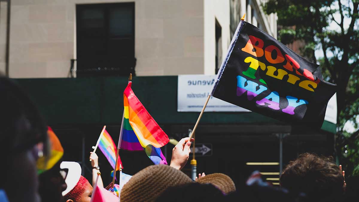 A crowd holds up rainbow flags at an LGBT Pride festival. One flag that says Born This Way and is held higher than the rest.