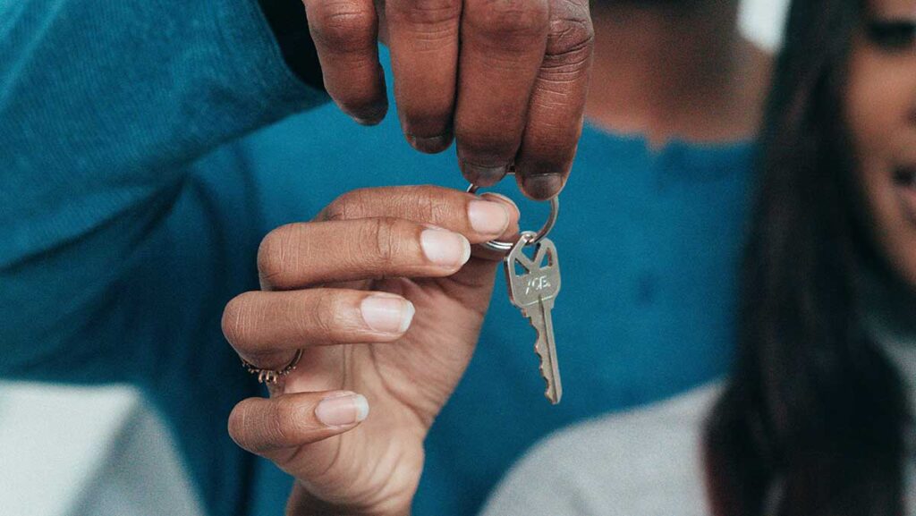 A female stands in front of a male. They're both holding a keyring with a house key attached. Their lender used interpreting serves to help them understand the house buying process.