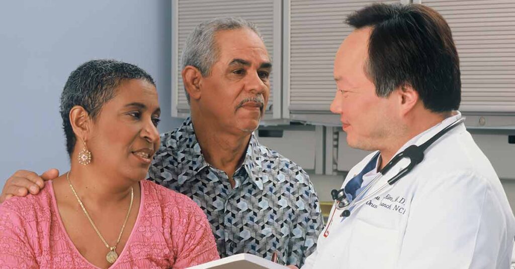 A Black woman and man with limited English proficiency talk with a male Asian doctor wearing a white lab coat and stethoscope, asking him for an interpreter.