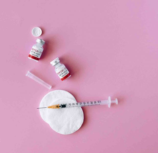 Two vials of the COVID-19 vaccine surrounded by a needle and cotton rounds on a pink background. 