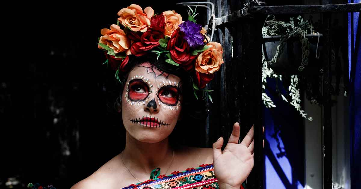 Female looking up. She's celebrating the Day of the Dead by wearing traditional Dia de los Muertos la Catrina makeup and a large flower crown on her head.