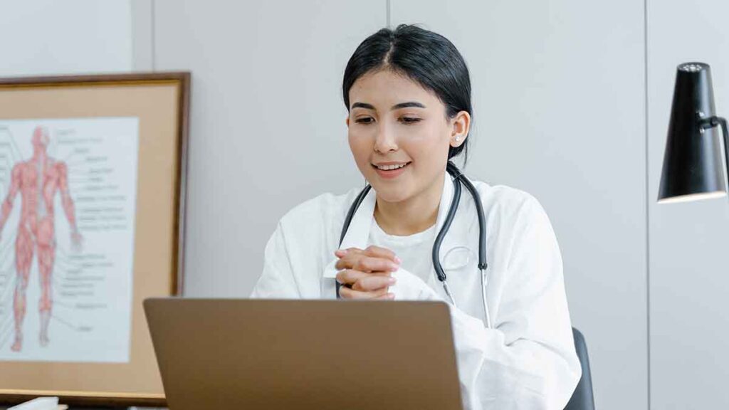 A female physician seeing a patient via a telehealth appointment. She's using a laptop and is wearing a white lab coat and stethoscope around her neck.