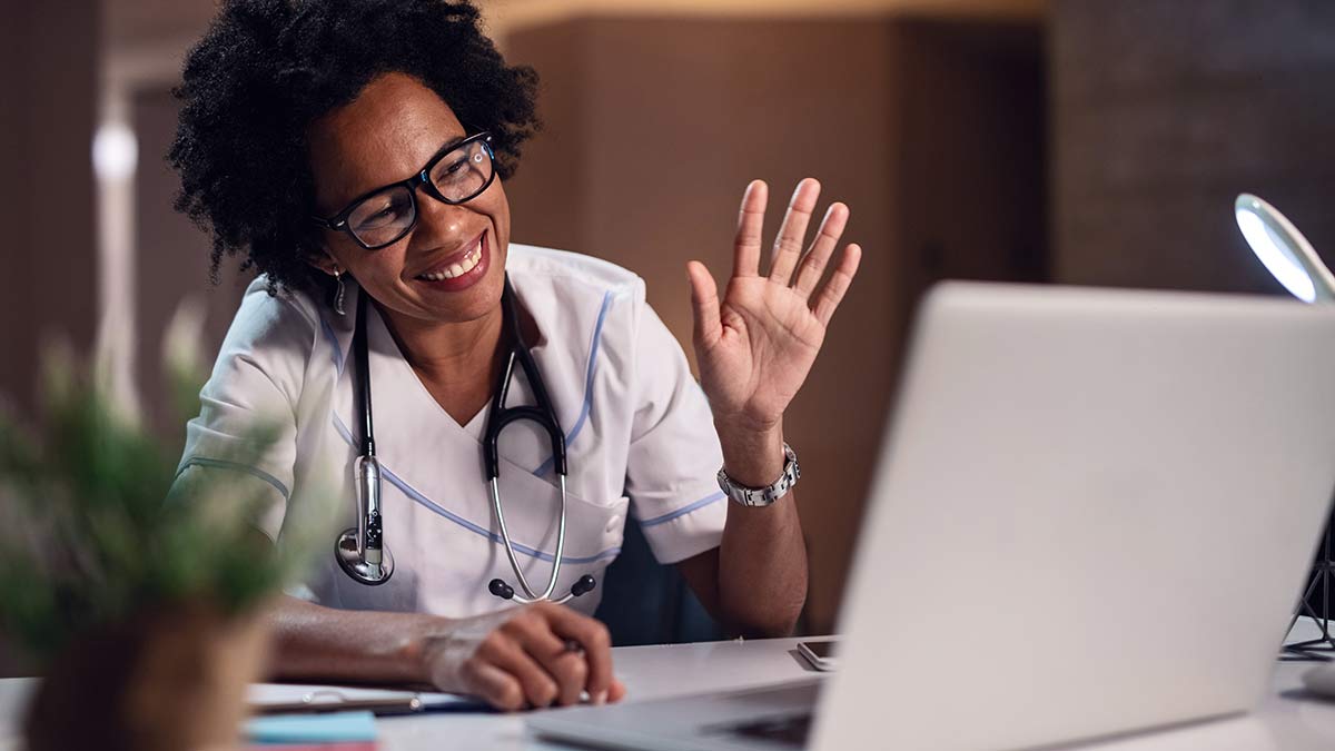 A doctor with a wide smile waves at a patient through a video call.