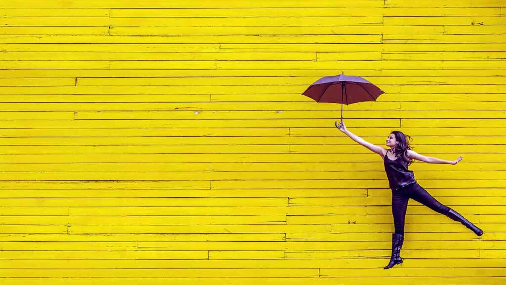 A woman holding an umbrella appears to float through the air in front of a wooden wall as she smiles and looks to the future.