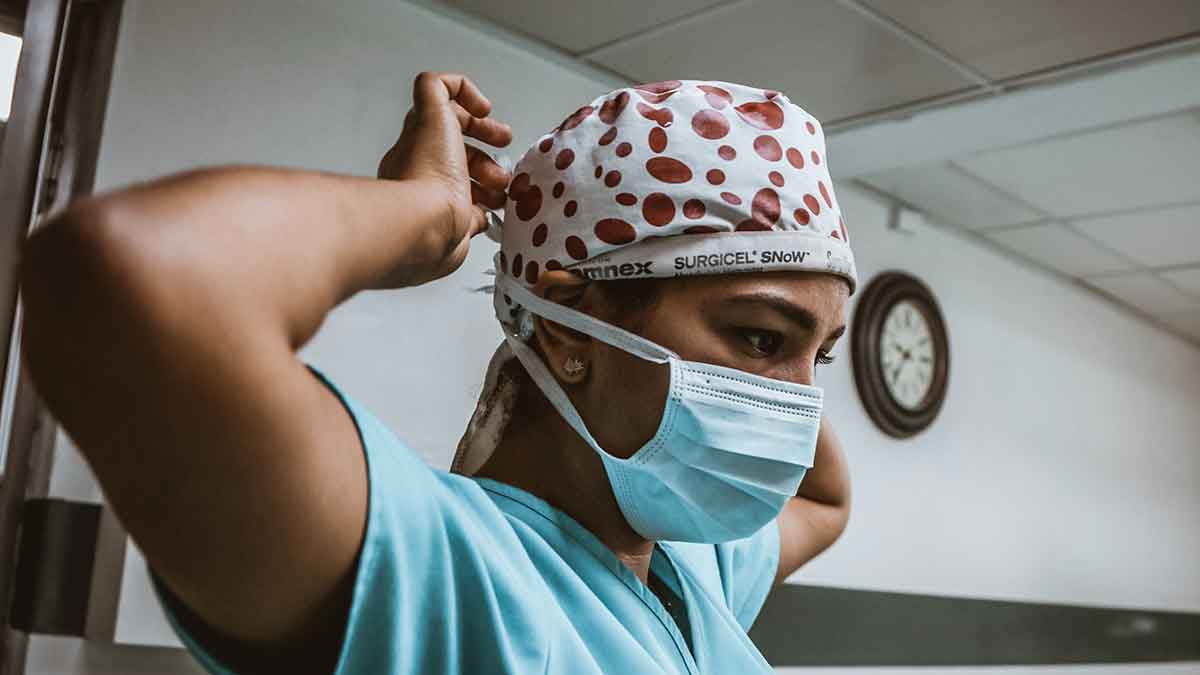 Female on-site healthcare interpreter ties a medical mask on her face. She's wearing scrubs and a white surgical cap with red dots.