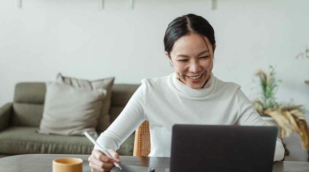 A female video remote interpreter smiles while sitting at her laptop. She's ready to take notes.