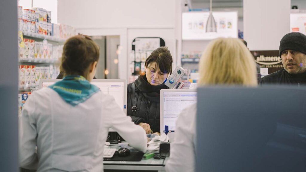 Two pharmacy staff members assist two customers at a checkout counter.