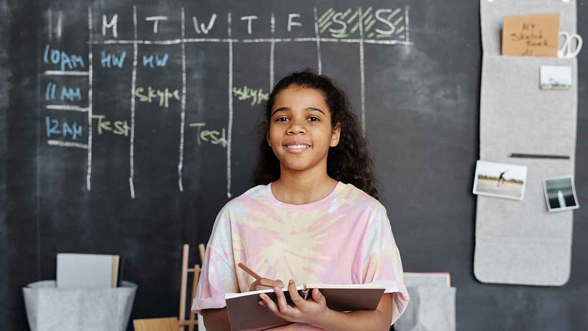 Smiling girl holding school notebook standing in front of blackboard.