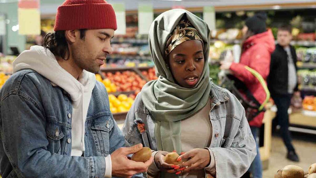 Man and woman both holding kiwis in mid-discussion at a grocery store. The man is wearing a red hat, sweatshirt, and denim jacket. The woman is wearing a mint headscarf and denim jacket.