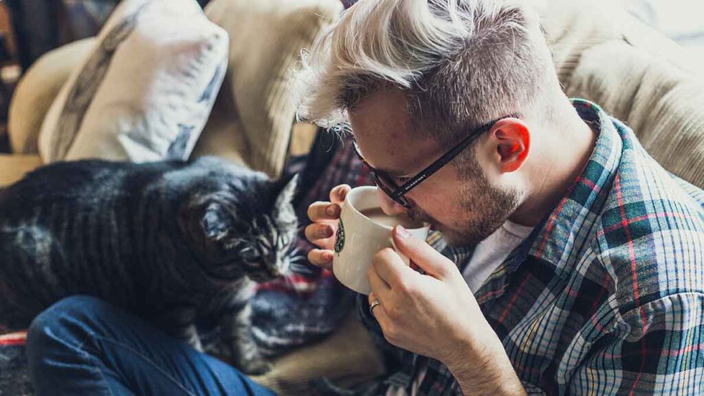 Video interpreter with glasses and a plaid shirt is sitting on his couch drinking coffee. His cat is beside him.