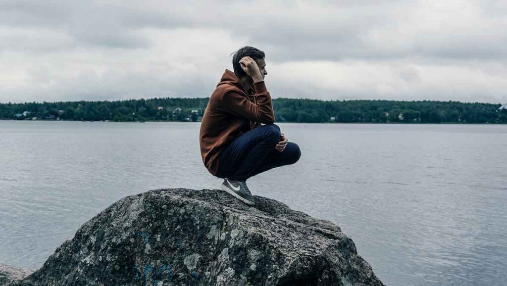 A young man squats on a giant rock overlooking the ocean. He's wearing a rust-colored sweatshirt and looks pensive.