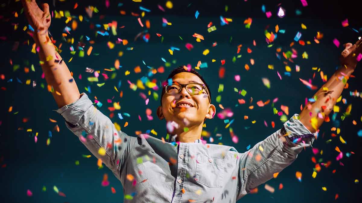 A grinning Asian man wearing glasses and a button-down shirt celebrates by throwing his arms into the air, releasing colorful confetti all around him.