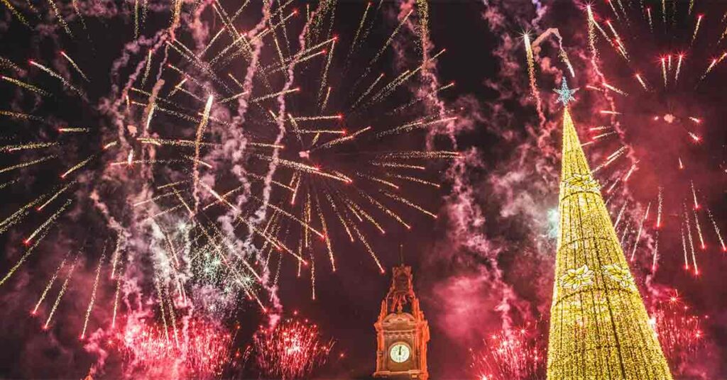 A firework display is set behind a tree of lights and a large clock tower that has struck midnight to celebrate Nochevieja.