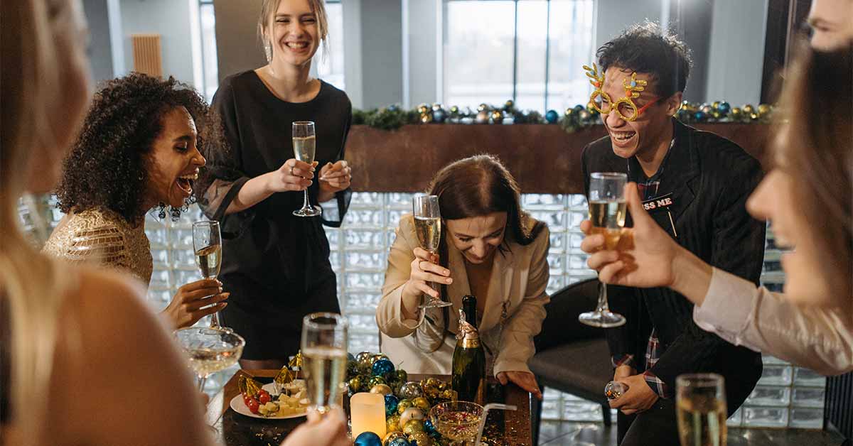 Seven friends laugh as they celebrate the new year together at home with party poppers, snacks, and champagne.