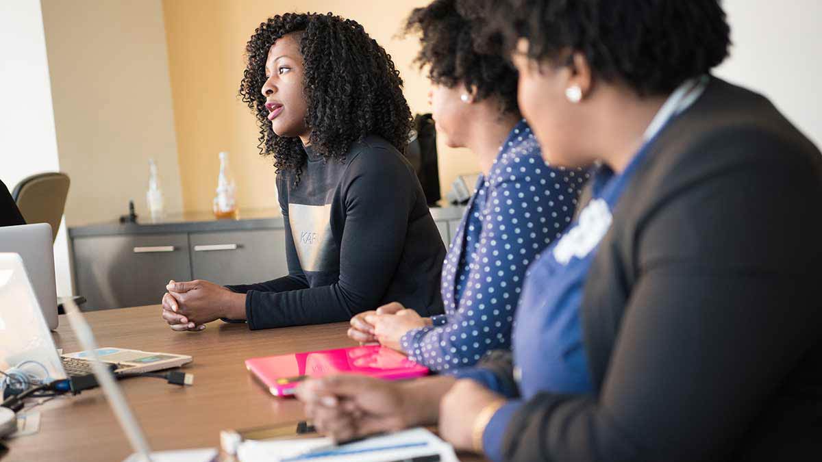 Three Black women interpreters with natural hair sit at a conference table talking about language access, with computers and documents in front of them.