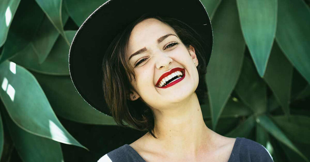 A smiling Latina woman, wearing a wide-brimmed black hat and red lipstick, stands in front of green leaves, happy that she got her interpreter certification.