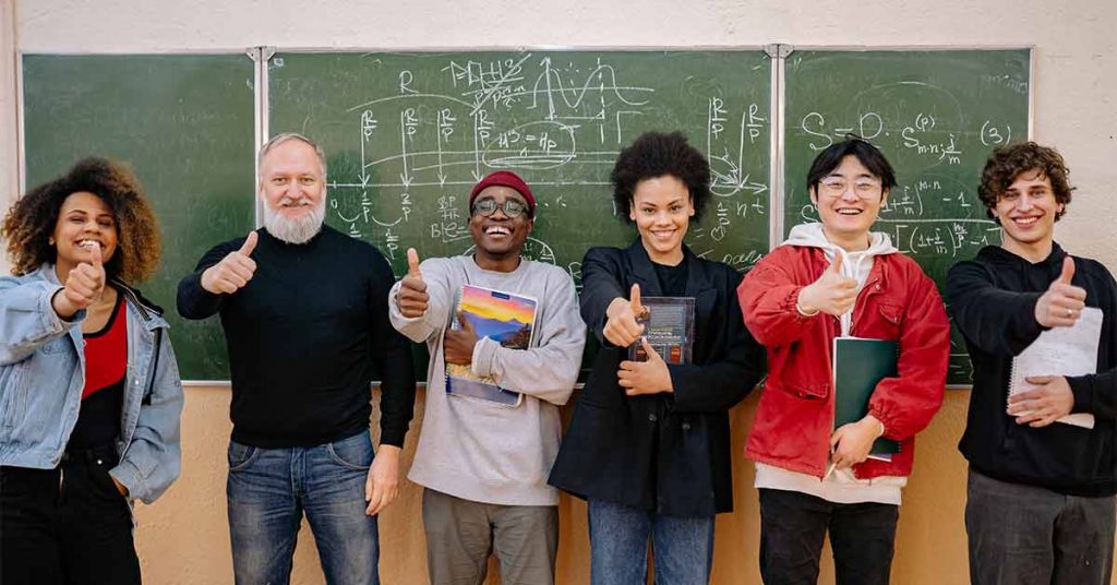 A professor and five students with big smiles and thumbs up stand in front of a chalkboard full of resettlement calculations.