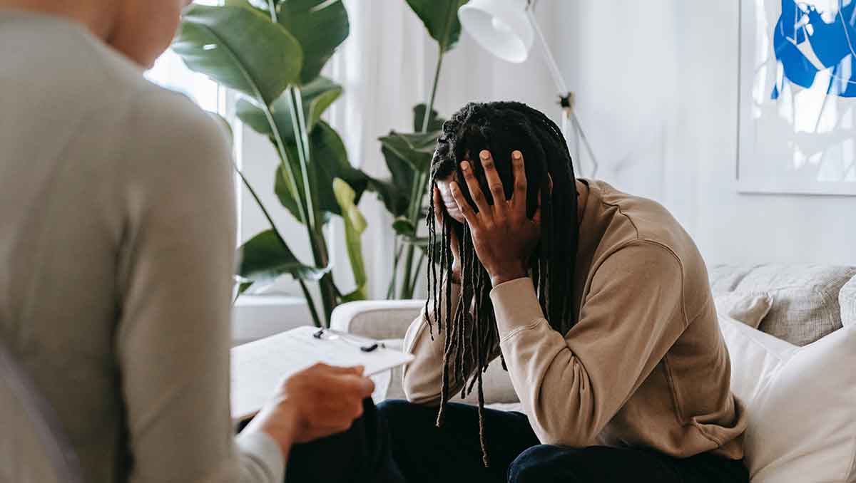 A man sits on a white couch with his head in his hands. A mental health provider sits facing him holding a clipboard.