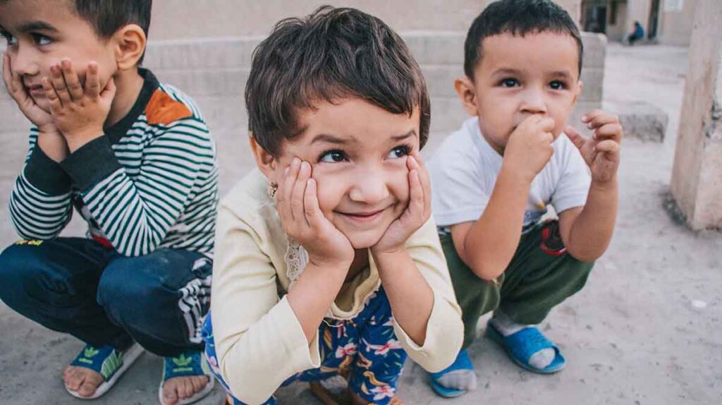 Three little boys crouch on the ground. The boy in front is smiling and resting his chin in his hands. The boy on the right has his hand to his mouth. The boy on the left is resting his chin in his hands.