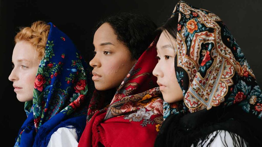 Three women wearing elaborately designed headscarves stand together and look into the distance with pensive expressions.