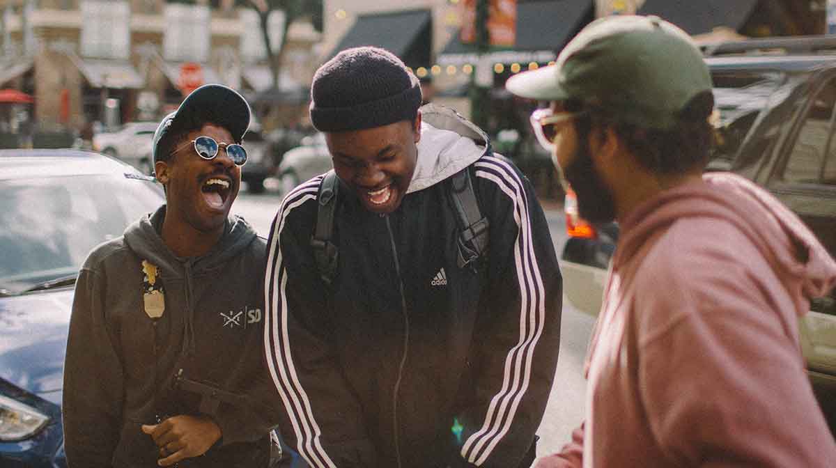 Three Black men talking outside by parked cars. All three are laughing.