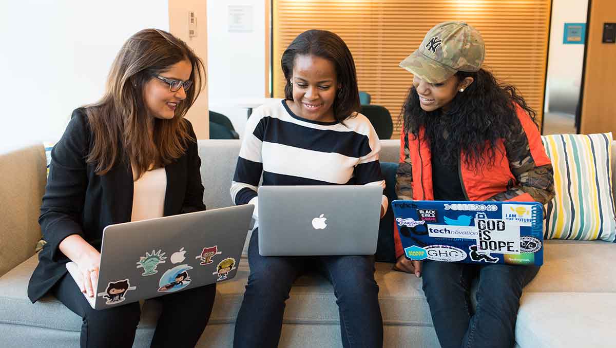 Three women sitting on a couch looking at their laptops. They are working together and smiling.