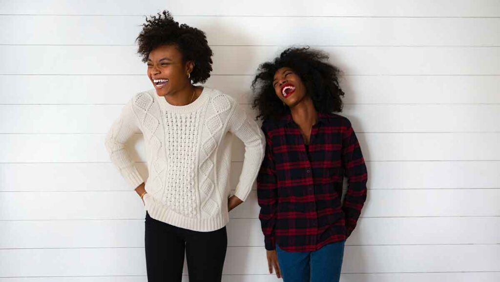 Two Black women stand against a white background. They are both laughing.