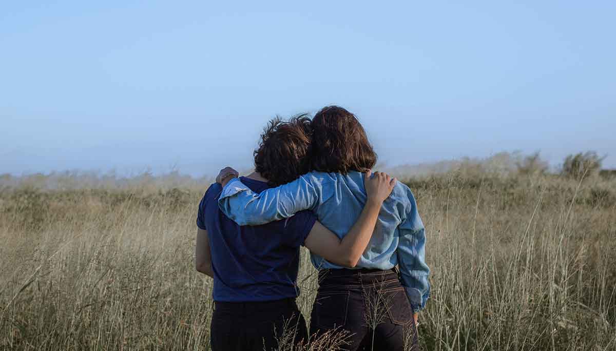 Two friends side hugging looking out over a field. One friend has their head on the other's shoulder.