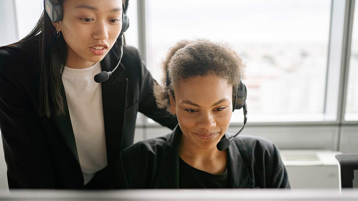 A call center manager coaches a call center agent who looks triumphantly at a computer monitor. They both wear headsets.