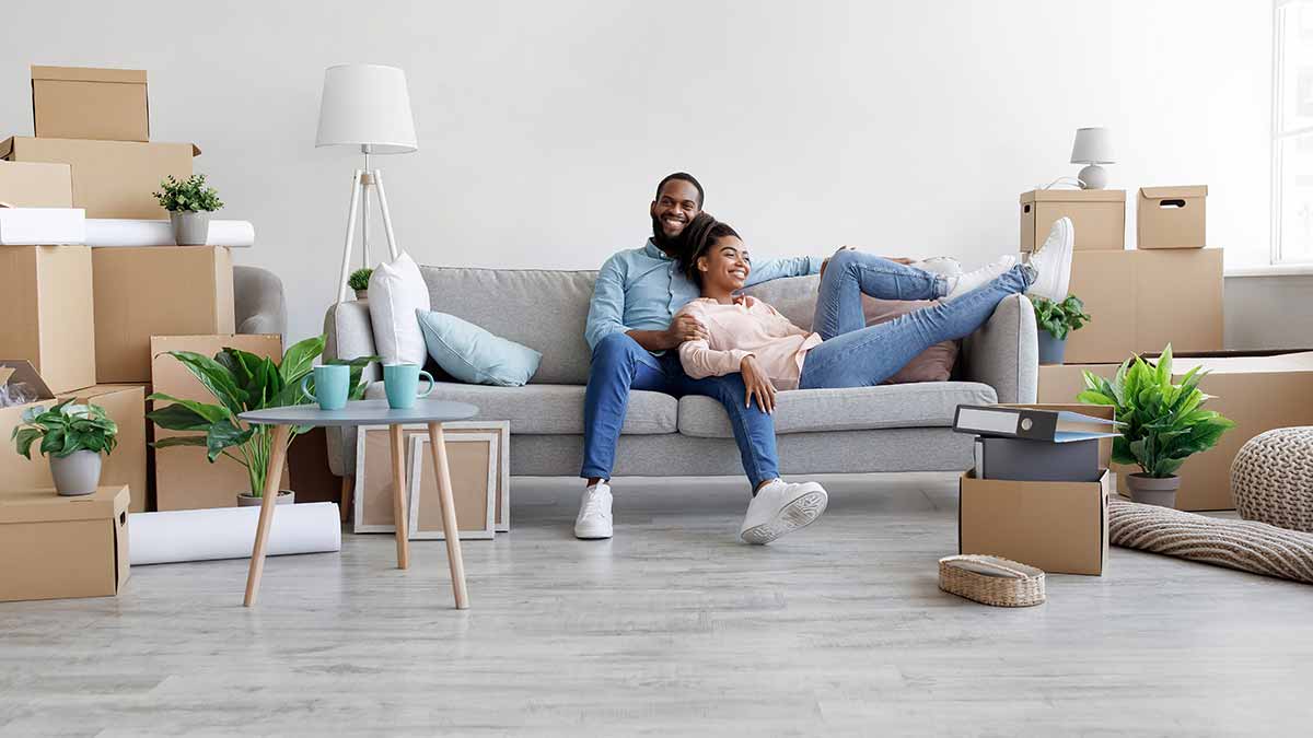 Two smiling homeowners sit on their new couch surrounded by moving boxes.