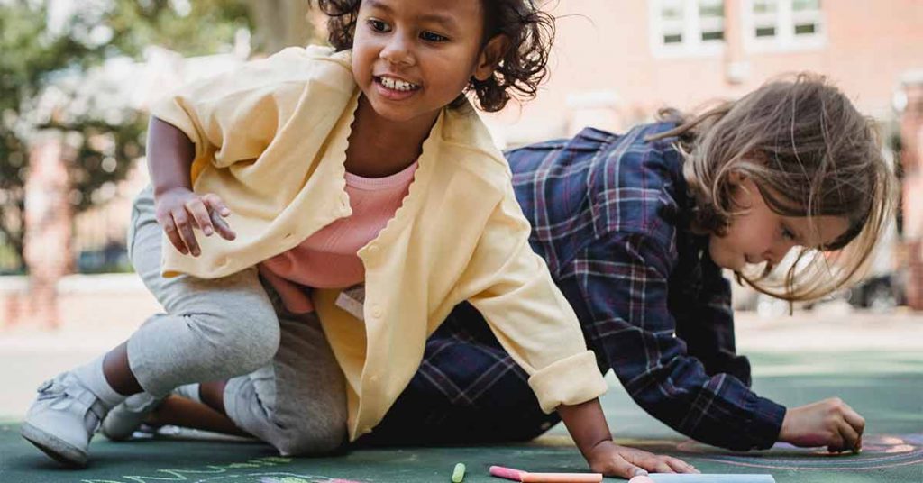 Two small children have fun drawing on the pavement with chalk together.