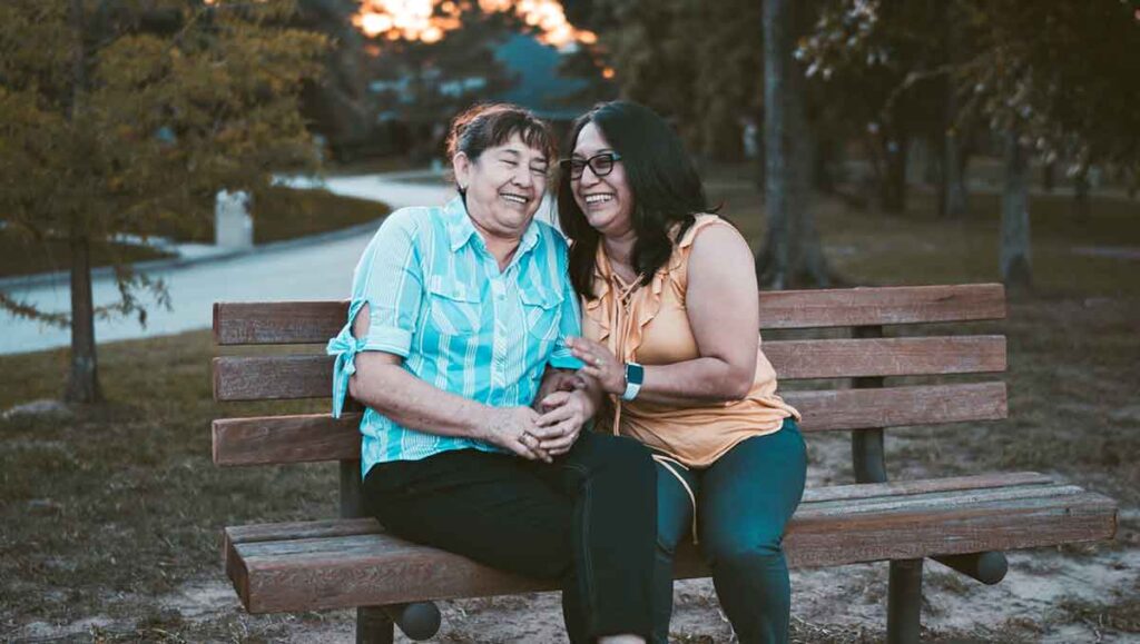 Two older women sit on a bench in a park. Both are laughing and having a great time. One is wearing a blue button-up shirt with her hair back. The other is wearing a peach tank top, a watch and glasses.