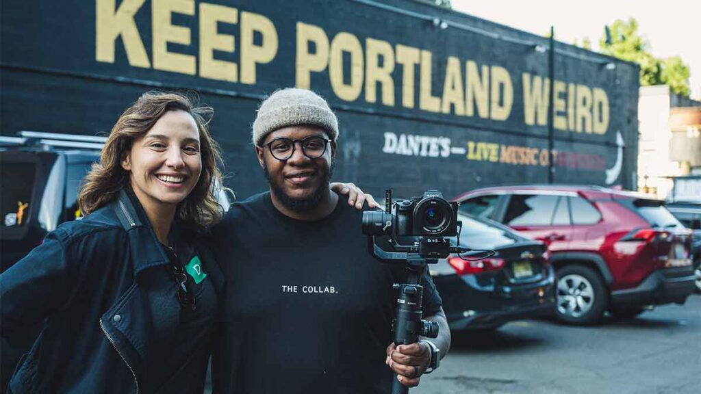 Two friends in Portland, Oregon stand with their arms around each other in front of a mural that says Keep Portland Weird.