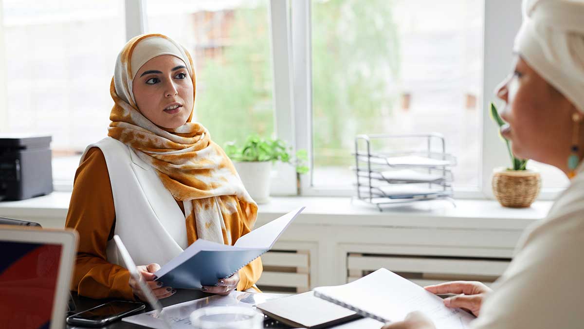 Two women sit at a table holding and discussing translated financial documents. One woman is wearing a yellow and white hijab.