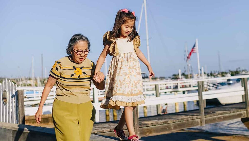 Older woman wearing yellow top and pants hold a young girl's hand