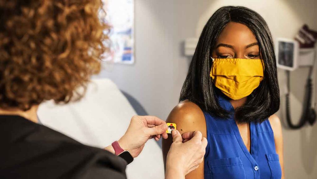 Black woman wearing a mask is getting a bandage applied to her arm after receiving a COVID-19 vaccine.