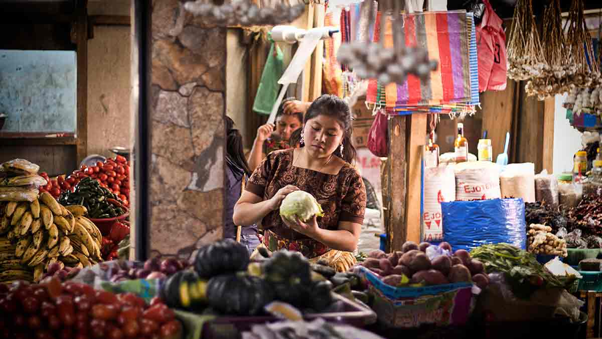 A woman looks at a head of lettuce in her hand at a market. She is surrounded by colorful bags and produce.