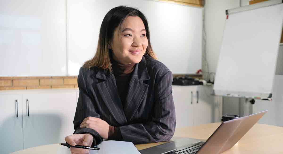 Smiling female video interpreter holding a pen and sitting at a table in front of a laptop.