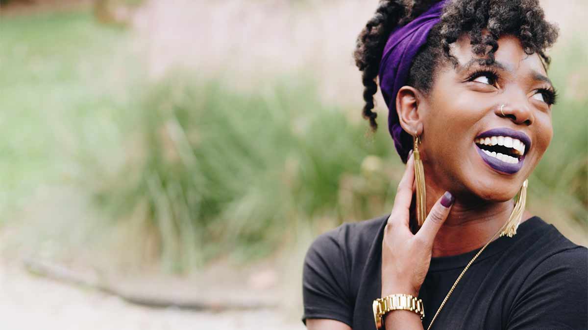 Black female smiling while looking over her shoulder. Her hand is touching her neck.