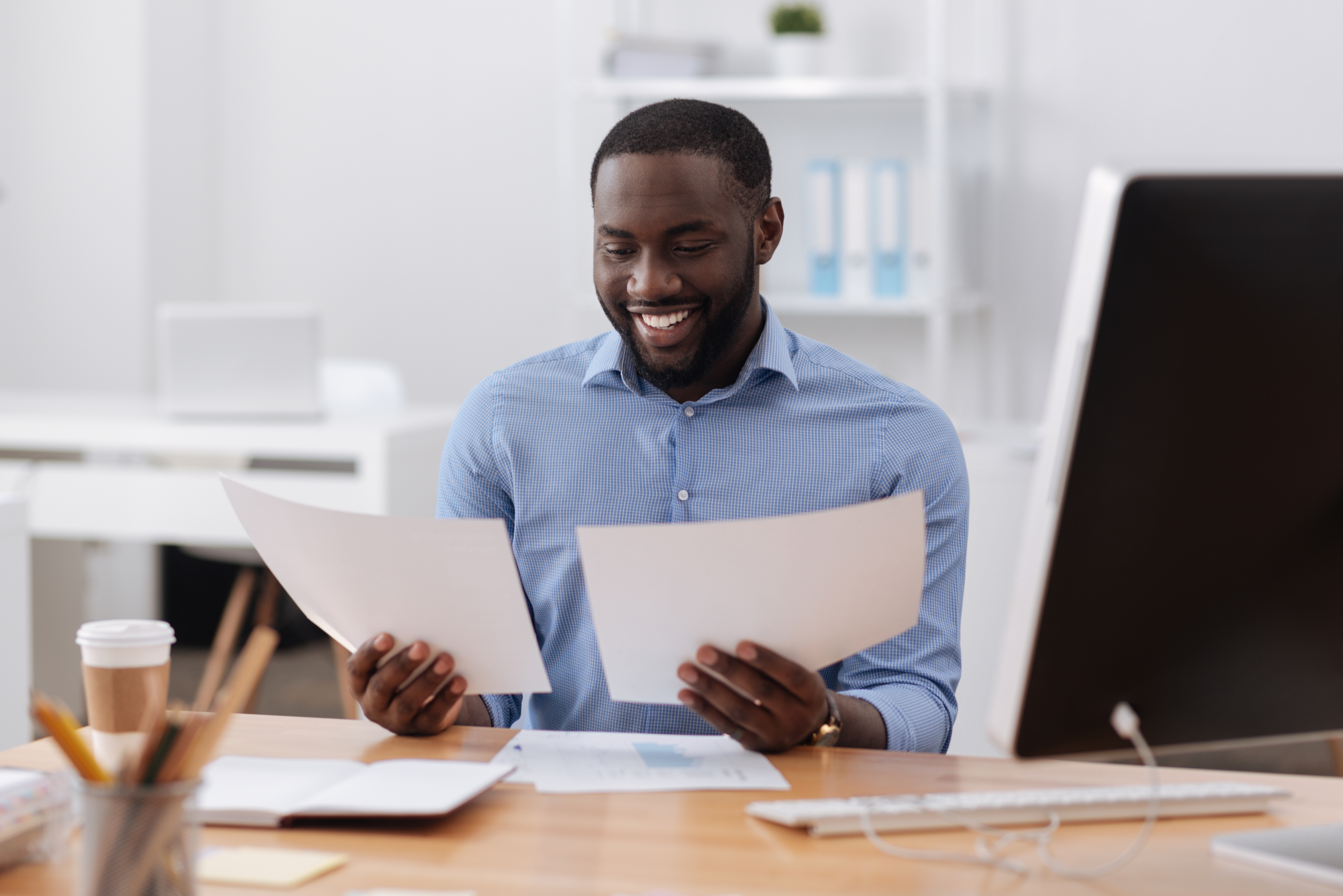 Man sitting at his desk smiling. He's holding and looking at two pieces of paper comparing qualified vs. certified interpreters.